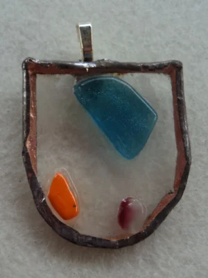 Pendentif en forme d'écusson en verre transparent. Un fragment bleu, un éclat orange et un rouges fondus en relief à la surface. Monté en Tiffany, une bélière soudée au dos.