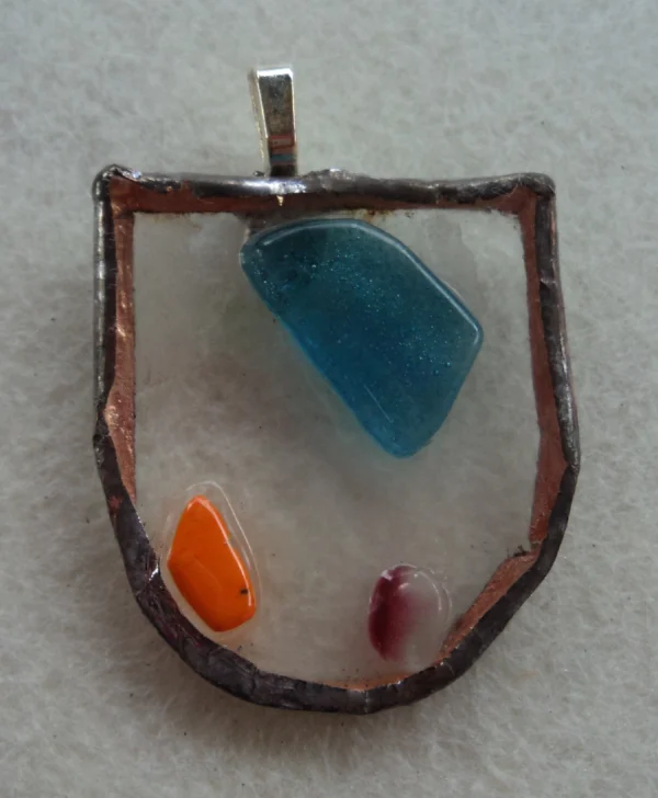 Pendentif en forme d'écusson en verre transparent. Un fragment bleu, un éclat orange et un rouges fondus en relief à la surface. Monté en Tiffany, une bélière soudée au dos.