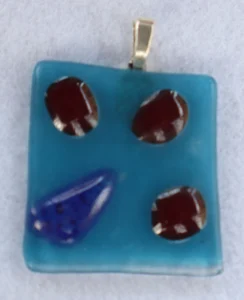 Pendentif carré en verre bleu avec 3 éclats rouge et un bleu fondus en relief à la surface. Une bélière argentée.