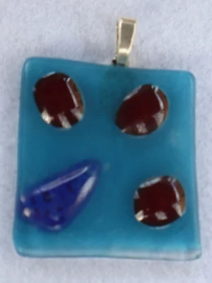 Pendentif carré en verre bleu avec 3 éclats rouge et un bleu fondus en relief à la surface. Une bélière argentée.