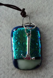 Pendentif en verre dichroïque bleu turquoise avec un tiret blanc u dessous, fondus sur du verre bleu nuit. Un anneau fondu entre 2 couches de verre a permis de fixer une breloque en forme de pioche