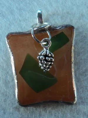 Pendentif en verre marron clair avec deux formes vertes fondues à la surface. Monté en Tiffany avec une bélière argentée, une petite pomme de pin y est suspendue