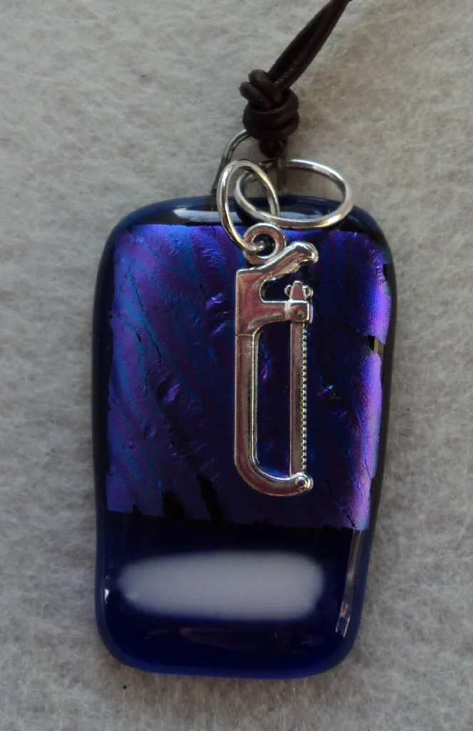 Pendentif en verre dichroïque bleu avec un tiret blanc. Un anneau a été inséré lors de la fusion. Un anneau en métal argenté permet de passer un pendentif en forme de scie.