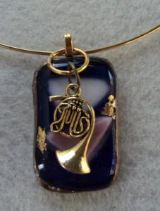 Pendentif en verre bleu avec 2 morceaux de verre de qualité Tiffany fondus et dépassant de la surface, Quelques éclats de feuille dorée ont été collés à la surface. Une bélière dorée soudée à l'arrière a permis de passer une breloque en forme de cor de chasse. Ce bijou est monté selon la technique Tiffany, serti de cuivre, soudé à l'étain et patiné en nuances de cuivre ancien. 