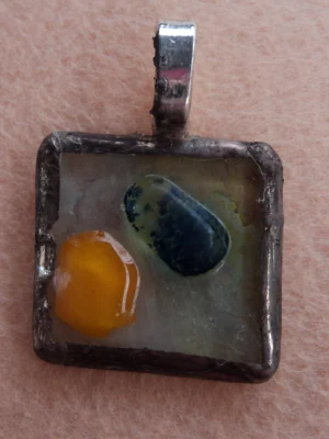 Petit pendentif carré en verre fondu avec des éclats orange et bleu. Monté en Tiffany, une bélière argentée.