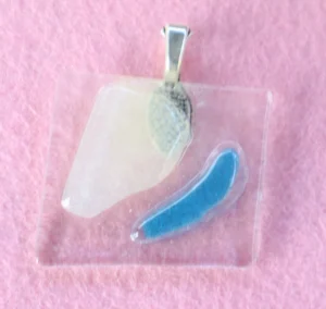 Pendentif en verre transparent sur lequel un éclat blanc et un bleu sont fondus. Une bélière en métal argentée est collée à l'arrière