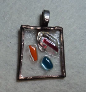 Pendentif rectangulaire en verre transparent, avec 3 éclats rouge, bleu et orange. Monté en Tiffany, serti de cuivre et soudé à l'étain, avec une patine cuivrée. Une bélière argentée soudée à l'arrière.