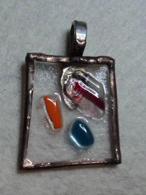 Pendentif rectangulaire en verre transparent, avec 3 éclats rouge, bleu et orange. Monté en Tiffany, serti de cuivre et soudé à l'étain, avec une patine cuivrée. Une bélière argentée soudée à l'arrière.