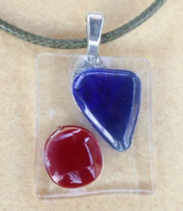 Pendentif rectangulaire en verre transparent avec un éclat triangulaire bleu et un cercle rouge. Une bélière argentée est collée à l'arrière