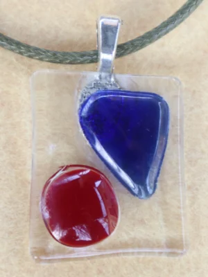 Pendentif rectangulaire en verre transparent avec un éclat triangulaire bleu et un cercle rouge. Une bélière argentée est collée à l'arrière