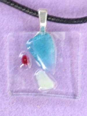 Pendentif rectangulaire en verre transparent, avec un éclat bleu, un blanc et un rouge plus petits. Une bélière en métal argenté est collée à l'arrière