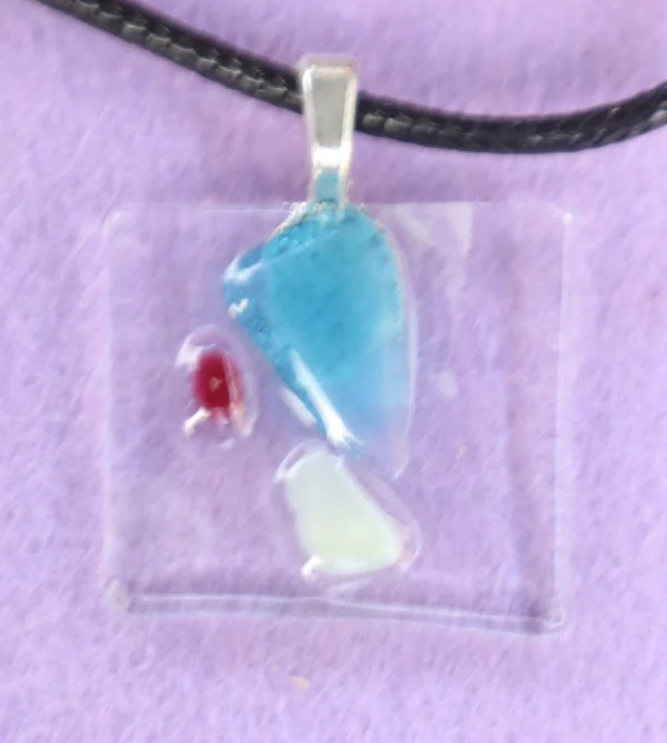 Pendentif rectangulaire en verre transparent, avec un éclat bleu, un blanc et un rouge plus petits. Une bélière en métal argenté est collée à l'arrière