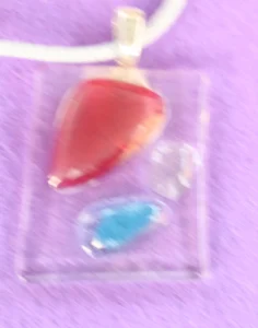 Pendentif rectangulaire en verre transparent avec 3 éclats triangulaires, un rouge plus gros que le bleu et le transparent. Une bélière argentée collée à l'arrière