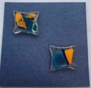 Carte postale carrée en carton bleu métallique. Deux carrés de verre, déformés par la fusion, ont été collés en diagonale. Ils sont parsemés d'éclats bleus et orange.