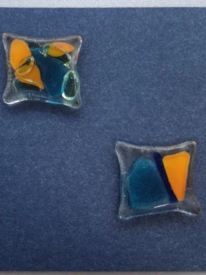 Carte postale carrée en carton bleu métallique. Deux carrés de verre, déformés par la fusion, ont été collés en diagonale. Ils sont parsemés d'éclats bleus et orange.