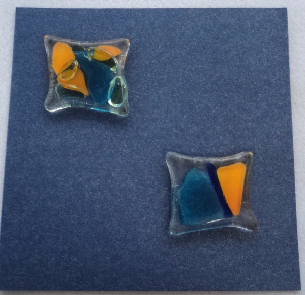 Carte postale carrée en carton bleu métallique. Deux carrés de verre, déformés par la fusion, ont été collés en diagonale. Ils sont parsemés d'éclats bleus et orange.