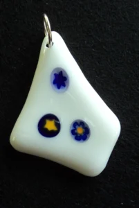 Pendentif en verre blanc en forme de losange irrégulier, avec 3 millefioris bleus, deux avec un cœur jaune. Un anneau argenté permet de le suspendre à une chaîne ou un lacet