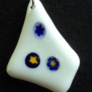 Pendentif en verre blanc en forme de losange irrégulier, avec 3 millefioris bleus, deux avec un cœur jaune. Un anneau argenté permet de le suspendre à une chaîne ou un lacet