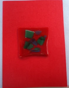 Carte postale rectangulaire en carton orange vermillon à 2 volets. Un carré de verre rouge intense est collé en son centre. Des éclats verts et bleus ont été fondus.