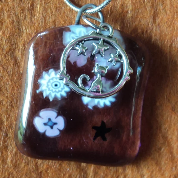 Pendentif en verre mauve semi transparent avec 6 millefioris blanc et rose. Une breloque représentant un chat assis dans un cercle sous des étoiles.