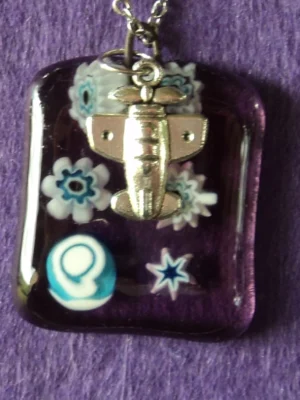 Pendentif rectangulaire en verre semi transparent mauve avec 6 millefioris blancs au cœur bleu pâle fondus. Une breloque en forme d'avion.