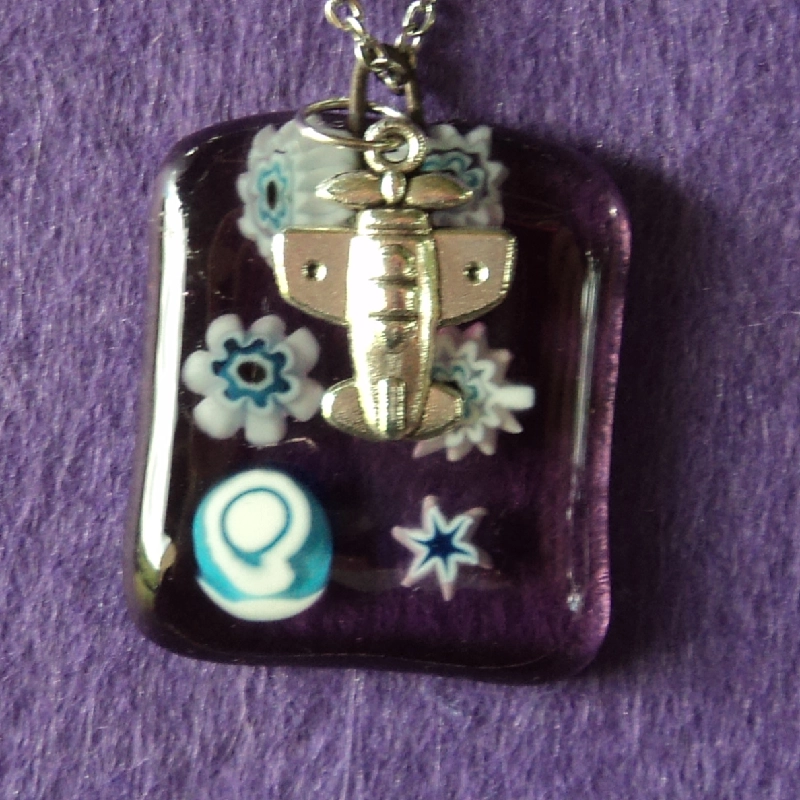 Pendentif rectangulaire en verre semi transparent mauve avec 6 millefioris blancs au cœur bleu pâle fondus. Une breloque en forme d'avion.