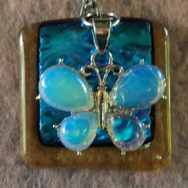 Pendentif carré en verre dichroïque bleu turquoise sur verre orange semi-transparent. Une breloque représentant un papillon en aigue-marine